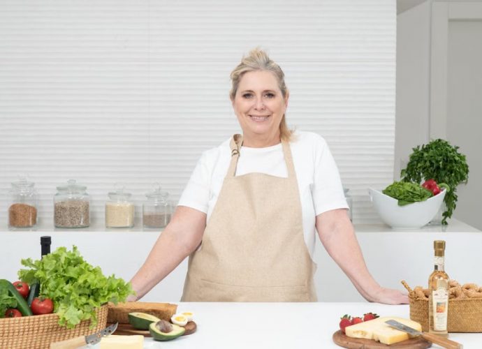 A mature woman in an apron stands at a kitchen counter with fresh produce and cheese, ready for cooking.
