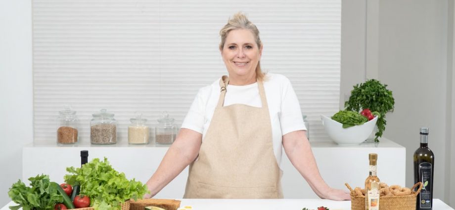A mature woman in an apron stands at a kitchen counter with fresh produce and cheese, ready for cooking.